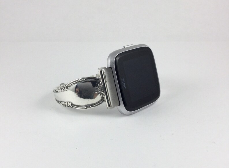 May include: A silver Fitbit smartwatch attached to a silver spoon bracelet. The watch face is black and displays the word "fitbit".