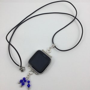 May include: A silver-toned Fitbit Versa smartwatch pendant necklace with a black band. The watch face is black. The pendant has a cluster of blue beads hanging from the bottom. The word "Fitbit" is visible on the watch face.