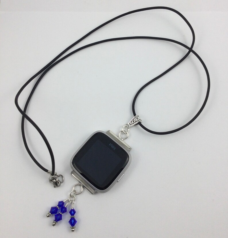 May include: A silver-toned Fitbit Versa smartwatch pendant necklace with a black band. The watch face is black. The pendant has a cluster of blue beads hanging from the bottom. The word "Fitbit" is visible on the watch face.
