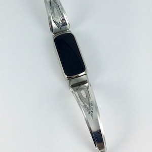 May include: A silver bracelet with a rectangular black stone center. The bracelet is made from repurposed silverware, with decorative etched patterns on the silver. The design is a unique blend of vintage and modern styles.