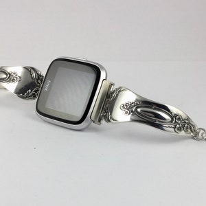 May include: Silver Fitbit smartwatch with a custom-made band crafted from a vintage silverware spoon. The band features intricate floral designs and a dangling charm.