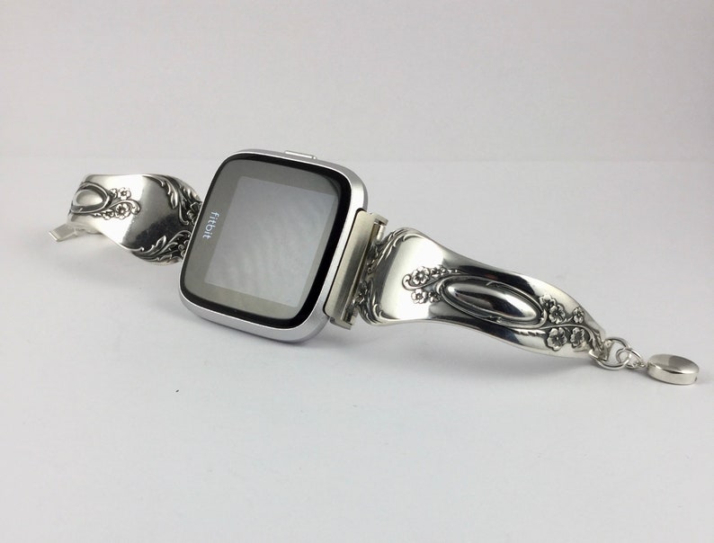 May include: Silver Fitbit smartwatch with a custom-made band crafted from a vintage silverware spoon. The band features intricate floral designs and a dangling charm.