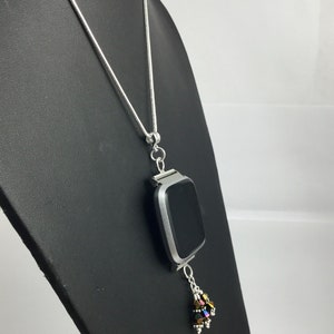 Puede incluir: Un collar plateado con un colgante rectangular, probablemente un reloj inteligente, con una pantalla negra. Un grupo de cuentas iridiscentes cuelga debajo. El collar se exhibe sobre un soporte negro sobre un fondo blanco.