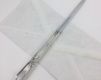 May include: A silver letter opener with an ornate handle featuring a floral design. The blade is sharp and pointed.