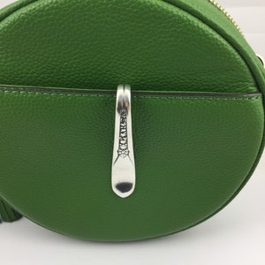 May include: A round, green leather handbag with a silver-colored spoon-shaped clasp. The bag has a gold zipper and a textured surface. The clasp is detailed with decorative patterns.