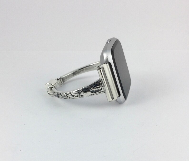 May include: A silver watch band with an ornate floral design. The watch face is square and black.