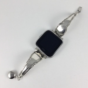 May include: Silver watch band made from a repurposed vintage spoon. The band is attached to a silver Fitbit smartwatch with a black screen. The band has a decorative design and a small silver charm.