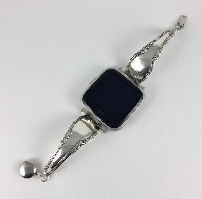 May include: Silver watch band made from a repurposed vintage spoon. The band is attached to a silver Fitbit smartwatch with a black screen. The band has a decorative design and a small silver charm.