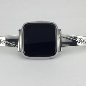 May include: A silver Fitbit Versa watch with a decorative silver bracelet band. The band has a floral design and a small charm.
