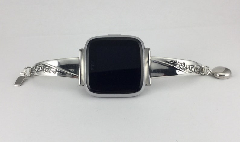 May include: A silver Fitbit Versa watch with a decorative silver bracelet band. The band has a floral design and a small charm.
