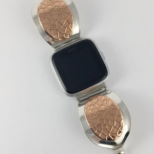 May include: A silver and rose gold watch band with a geometric pattern. The band is designed to fit a Fitbit watch. The watch face is black and displays the word "Fitbit".