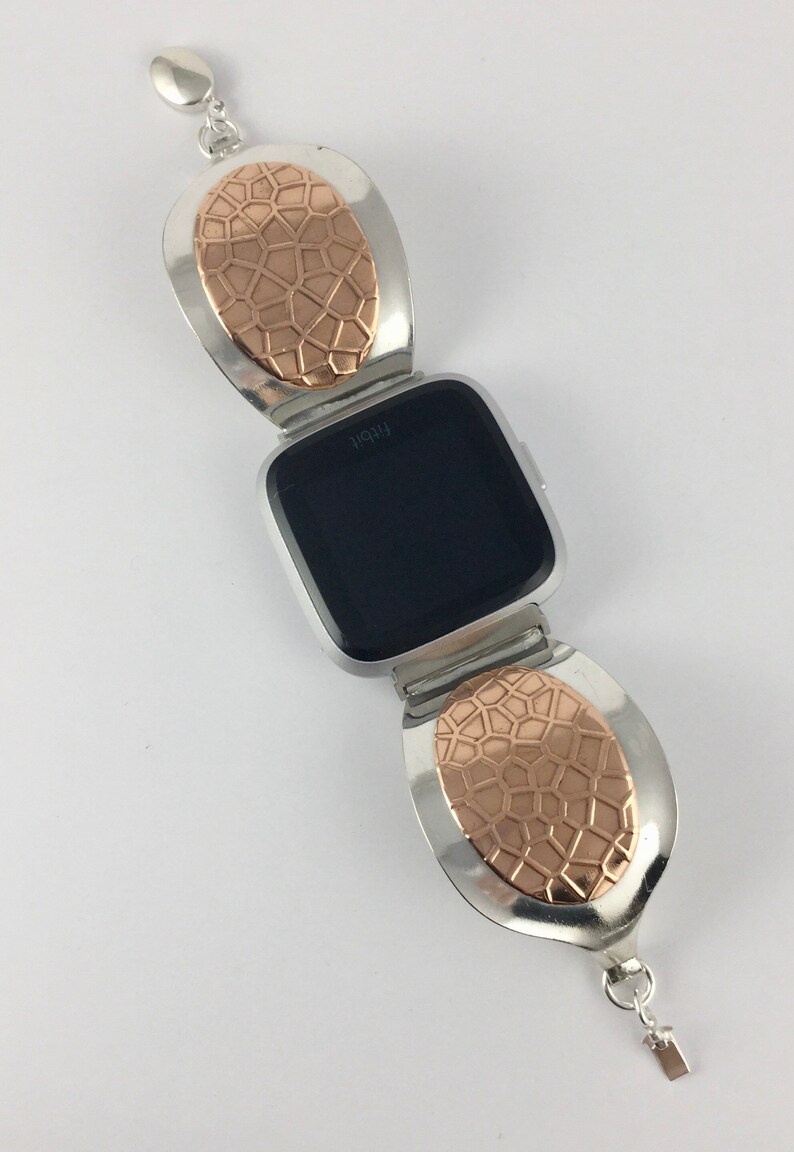 May include: A silver and rose gold watch band with a geometric pattern. The band is designed to fit a Fitbit watch. The watch face is black and displays the word "Fitbit".