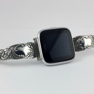 Puede incluir: Una pulsera de reloj plateada con un dise&ntilde;o adornado. La pulsera est&aacute; hecha de metal y tiene un patr&oacute;n floral. La pulsera de reloj es compatible con un reloj Fitbit.