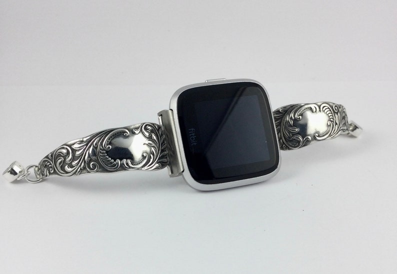 Puede incluir: Una pulsera de reloj plateada con un dise&ntilde;o adornado. La pulsera est&aacute; hecha de metal y tiene un patr&oacute;n floral. La pulsera de reloj es compatible con un reloj Fitbit.