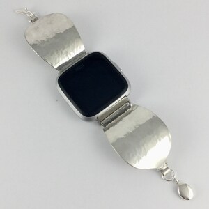 May include: A silver watch band with a hammered metal design. The band is made of two large, rounded pieces that fold over the watch face. The watch face is black and has the word "Fitbit" printed on it.