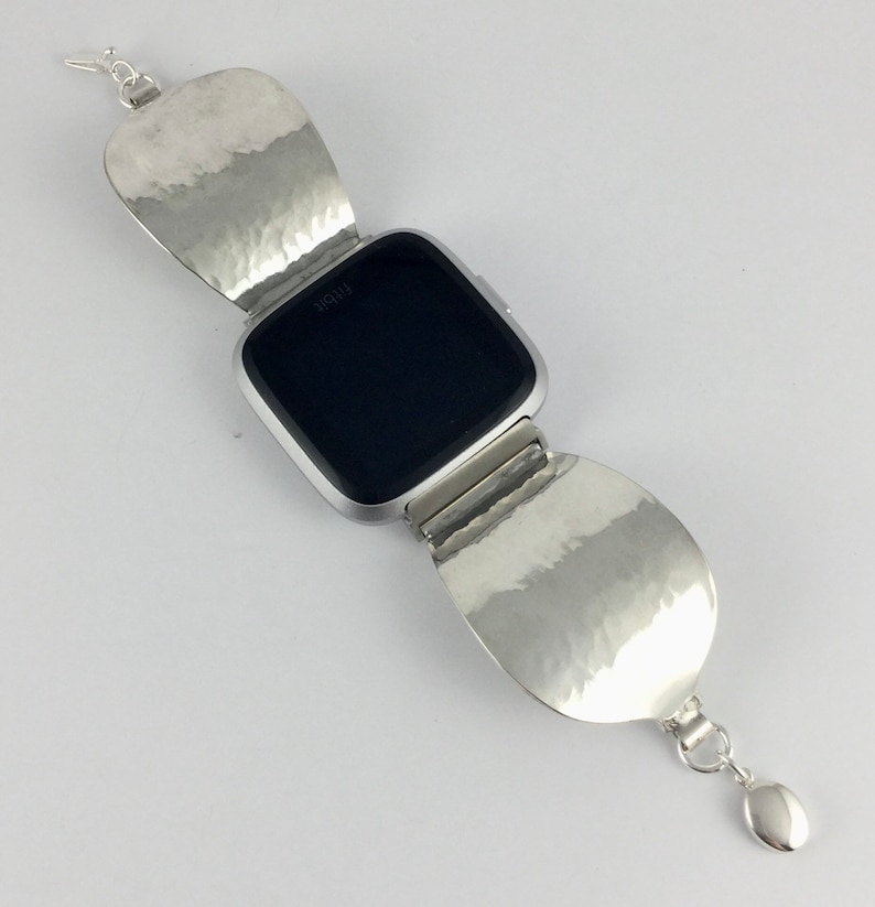 May include: A silver watch band with a hammered metal design. The band is made of two large, rounded pieces that fold over the watch face. The watch face is black and has the word "Fitbit" printed on it.