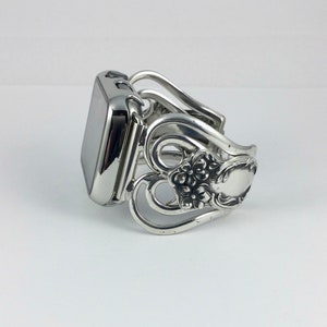 May include: A silver-toned Apple Watch band crafted from repurposed silverware. The band features ornate floral and scrollwork designs, with a polished finish. The watch face is a dark, reflective color, contrasting with the bright band.