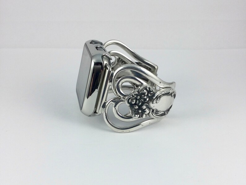 May include: A silver-toned Apple Watch band crafted from repurposed silverware. The band features ornate floral and scrollwork designs, with a polished finish. The watch face is a dark, reflective color, contrasting with the bright band.