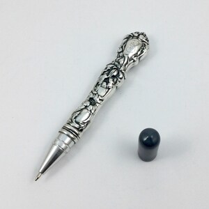 May include: Ornate silver pen with floral and scroll detailing. The pen has a cap and a fine point. The pen is a vintage style and is likely a collectible item. The pen is on a white background.