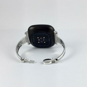 Puede incluir: Un brazalete plateado con un reloj Fitbit negro adjunto. La esfera del reloj es visible y muestra el logotipo de Fitbit y las letras "CE".