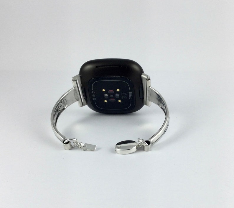 Puede incluir: Un brazalete plateado con un reloj Fitbit negro adjunto. La esfera del reloj es visible y muestra el logotipo de Fitbit y las letras "CE".
