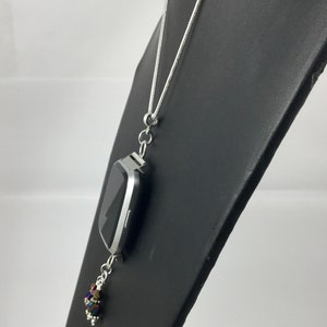 Puede incluir: Collar plateado con un colgante de reloj inteligente rectangular. El reloj tiene una pantalla negra y cuelga de una cadena fina. Un grupo de cuentas iridiscentes cuelga debajo, agregando un toque de color.