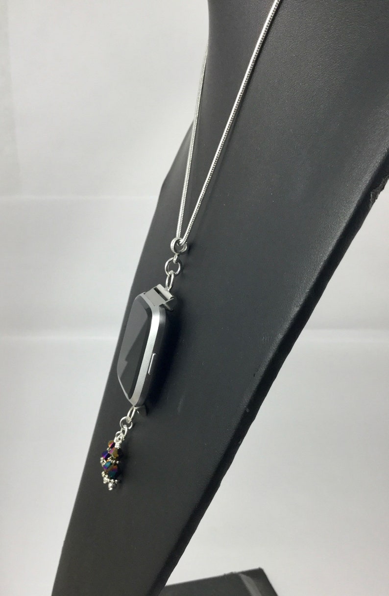 Puede incluir: Collar plateado con un colgante de reloj inteligente rectangular. El reloj tiene una pantalla negra y cuelga de una cadena fina. Un grupo de cuentas iridiscentes cuelga debajo, agregando un toque de color.