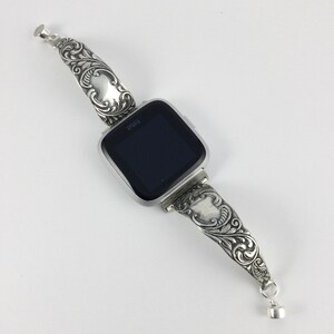 Puede incluir: Una pulsera de reloj de plata con un intrincado dise&ntilde;o floral. La pulsera est&aacute; unida a un reloj inteligente de forma cuadrada con una pantalla negra.