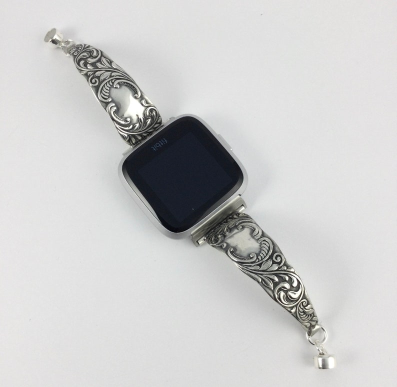 Puede incluir: Una pulsera de reloj de plata con un intrincado dise&ntilde;o floral. La pulsera est&aacute; unida a un reloj inteligente de forma cuadrada con una pantalla negra.