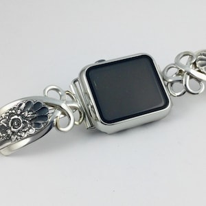 May include: A silver-toned smartwatch with a rectangular black screen, attached to a decorative metal watch band featuring intricate floral relief patterns and looped, open-work silver links on both sides of the watch casing.