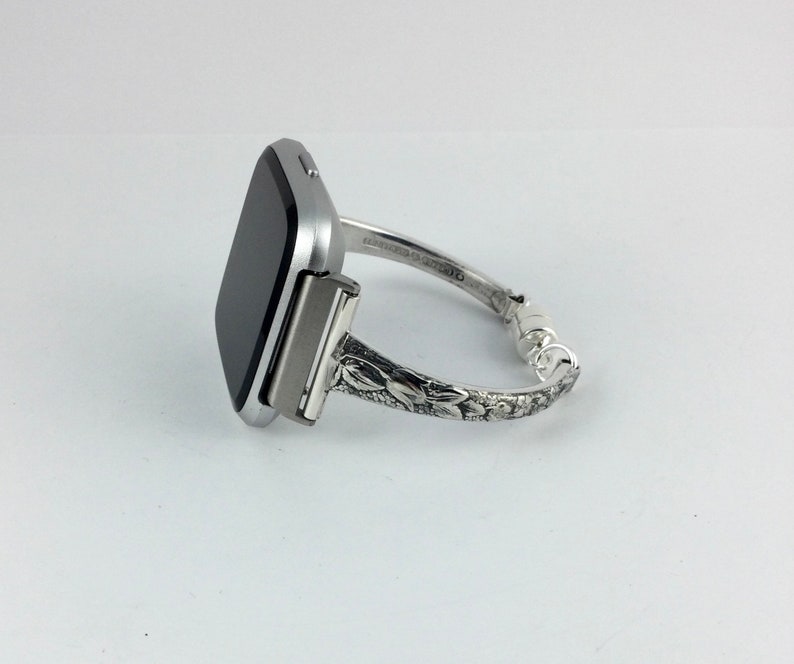 May include: A silver smartwatch with a black screen is attached to a silver bracelet with an ornate floral design.