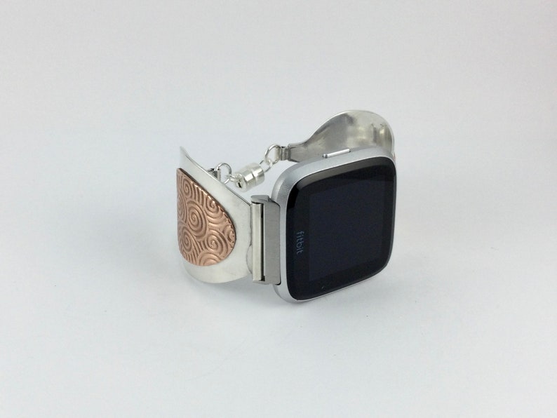 May include: A silver and rose gold metal watch band with a swirl pattern on the clasp. The band is designed to fit a Fitbit watch.