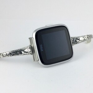 May include: A silver Fitbit smartwatch with a floral patterned band. The band is attached to the watch with a silver clasp.