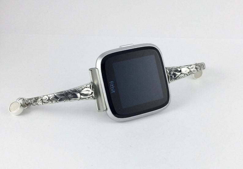 May include: A silver Fitbit smartwatch with a floral patterned band. The band is attached to the watch with a silver clasp.