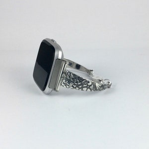 May include: Silver watch band with a floral design. The band is compatible with a square-shaped smartwatch.