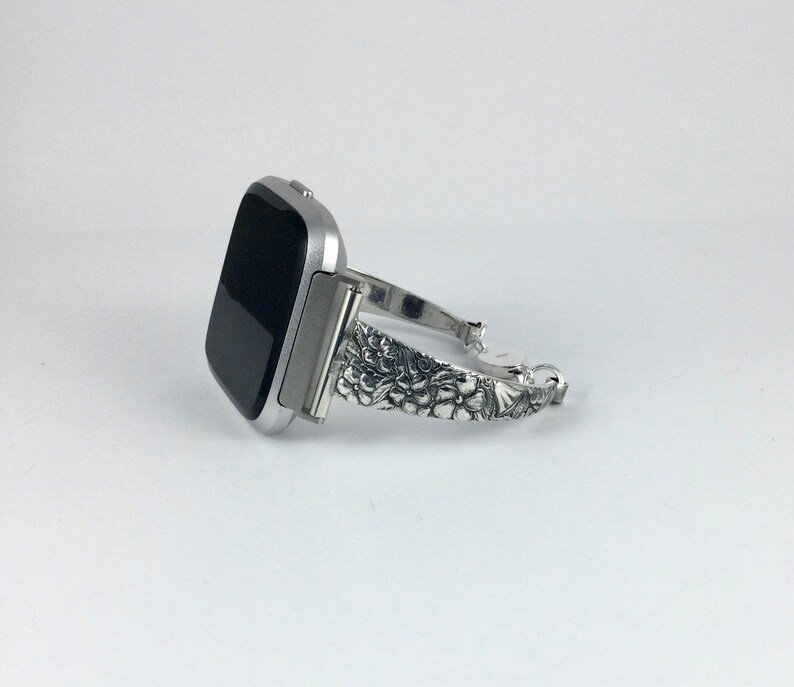 May include: Silver watch band with a floral design. The band is compatible with a square-shaped smartwatch.