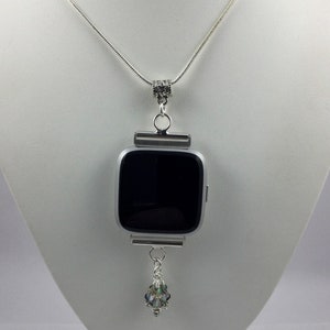 K&ouml;nnte beinhalten: Eine silberne Kettenkette mit einem silbernen Anh&auml;nger, der eine quadratische Smartwatch h&auml;lt. Die Smartwatch hat einen schwarzen Bildschirm und ein silbernes Armband. Ein kleiner Kristallanh&auml;nger h&auml;ngt am unteren Ende des Anh&auml;ngers.
