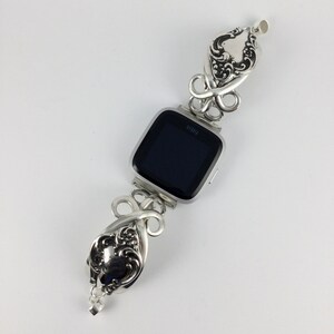 May include: Silver watch band made from repurposed silverware with a decorative floral design. The band is attached to a black-faced smartwatch with the word "Fitbit" printed on the face.