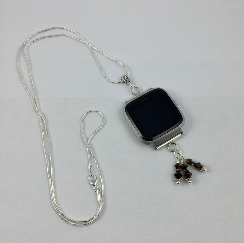Puede incluir: Un collar plateado con un colgante dise&ntilde;ado para sujetar un reloj Fitbit. La esfera del reloj es negra y el colgante tiene un adorno decorativo de cuentas marrones. El collar est&aacute; hecho de una fina cadena de serpiente.