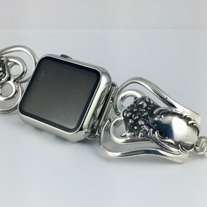 May include: Silver-toned Apple Watch band with a decorative floral design. The band is made from repurposed silverware, featuring ornate details and a vintage aesthetic. The watch face is square and has a black screen.