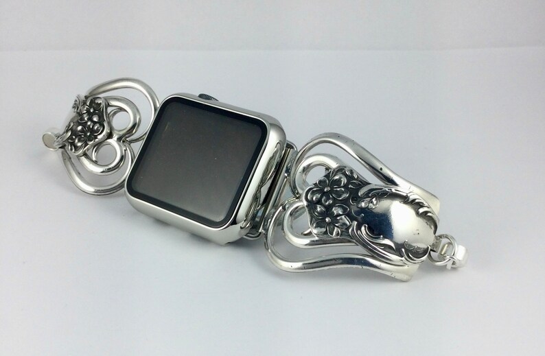 May include: Silver-toned Apple Watch band with a decorative floral design. The band is made from repurposed silverware, featuring ornate details and a vintage aesthetic. The watch face is square and has a black screen.