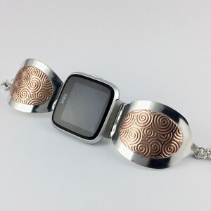 May include: A silver bracelet with a Fitbit watch attached. The bracelet has two oval-shaped pieces with a swirling copper design.