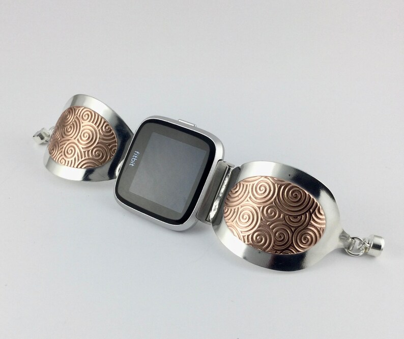 May include: A silver bracelet with a Fitbit watch attached. The bracelet has two oval-shaped pieces with a swirling copper design.