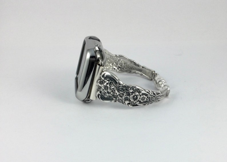 May include: A silver-toned Apple Watch case and band. The band features an ornate floral design, with detailed carvings and a polished finish. The watch face is a dark color, and the band appears to be made of metal.