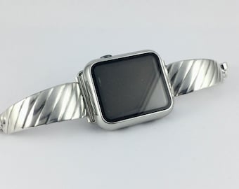 Puede incluir: Un reloj inteligente de tono plateado con una pantalla negra rectangular y una correa metálica con textura acanalada diagonal. La caja del reloj es de plata pulida y la correa consta de dos segmentos simétricos unidos a la unidad central.