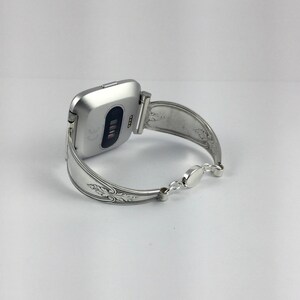 May include: Silver watch band made from a repurposed spoon with an ornate design. The band is attached to a silver smartwatch with a black screen.