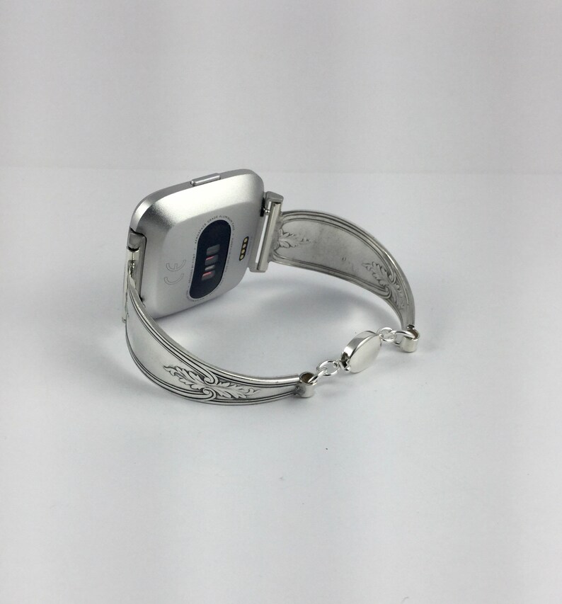 May include: Silver watch band made from a repurposed spoon with an ornate design. The band is attached to a silver smartwatch with a black screen.
