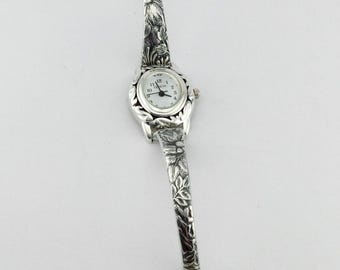Könnte beinhalten: Eine silberfarbene Armbanduhr mit einem ovalen weißen Zifferblatt, schwarzen Ziffern und dem Schriftzug Geneva. Das Armband ist durchgehend mit einem detaillierten floralen Prägemuster versehen und besitzt einen kleinen runden Verschluss.