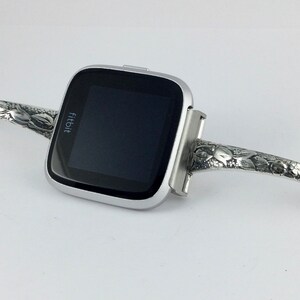 May include: A silver Fitbit smartwatch with a floral patterned band. The band is made of metal and has a clasp closure.