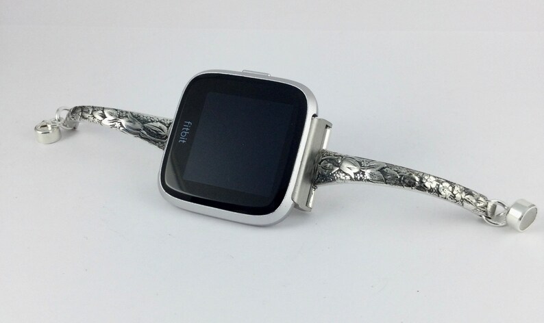 May include: A silver Fitbit smartwatch with a floral patterned band. The band is made of metal and has a clasp closure.